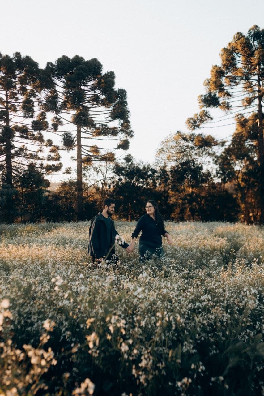 Camila e Bernardo caminhando de mãos dadas entre flores brancas